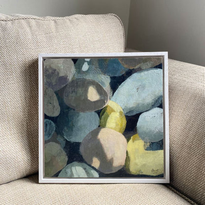 A framed square painting of smooth pebbles in muted blue, gray, beige, and yellow tones, leaning against a beige upholstered armchair.