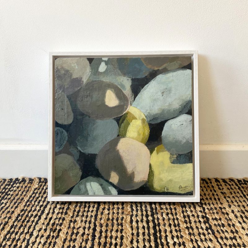 A framed square painting of smooth stones in muted gray, blue-gray, beige, and yellow-green tones, leaning against a white wall on a woven rug.